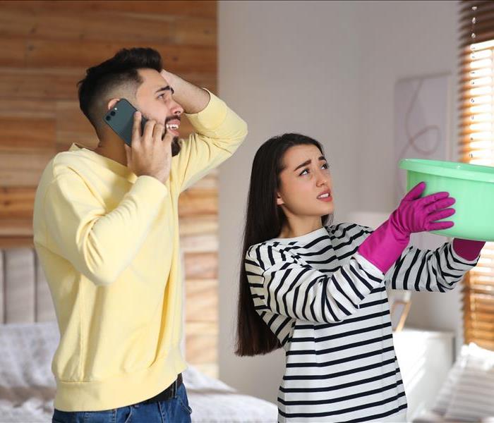 Young woman collecting leaking water from ceiling while her boyfriend calling roof repair service at home